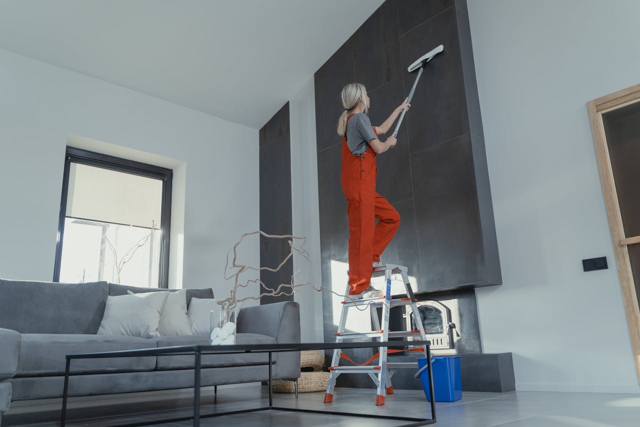 hero-img-02 A woman stands on a ladder in a modern living room, cleaning the high wall with a mop.